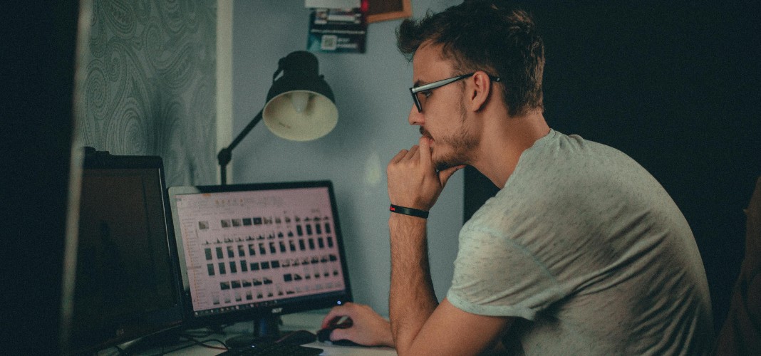 A man wearing glasses is sitting at a desk, looking intently at a computer screen displaying a grid of images. He is resting his chin on his hand, appearing focused. On the desk, there is a second monitor and a desk lamp. The room is dimly lit, creating a concentrated atmosphere.