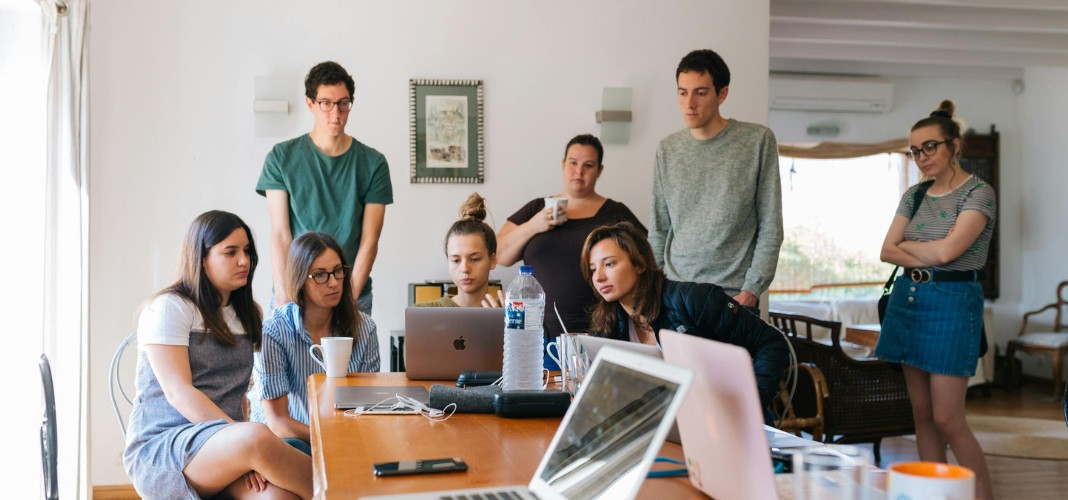 A group of eight people are gathered around a wooden table in a room illuminated by natural light. They are attentively looking at a laptop. On the table, there are coffee cups, a water bottle, and other items. Some of the people are seated, while others are standing. Everyone appears focused and engaged in what they are viewing on the screen.